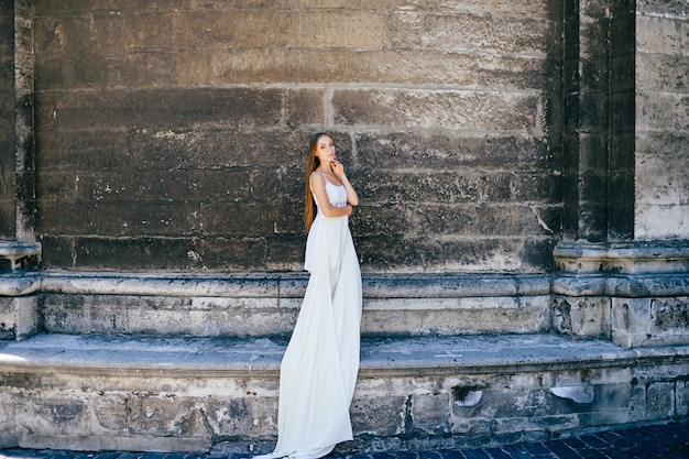 사진 돌 고대 벽 위에 포즈를 취하는 긴 흰 드레스에 젊은 낭만적 인 우아한 소녀