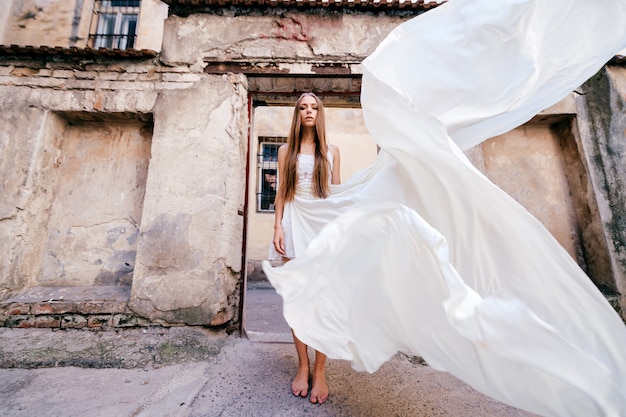 Молодая романтическая элегантная девушка в длинном летящем белом платье позирует над каменными древними зданиями