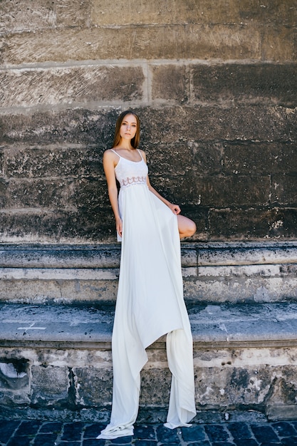 Foto giovane ragazza elegante romantica in vestito bianco lungo che posa sopra il muro antico di pietra