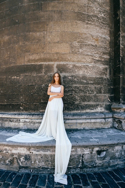 Giovane ragazza elegante romantica in vestito bianco lungo che posa sopra il muro antico di pietra