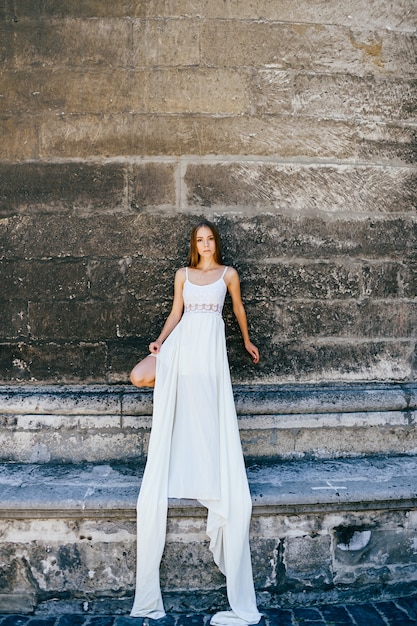 Foto giovane ragazza elegante romantica in vestito bianco lungo che posa sopra il muro antico di pietra