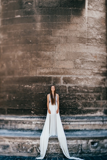 Giovane ragazza elegante romantica in vestito fluido bianco lungo che posa sopra il muro antico di pietra