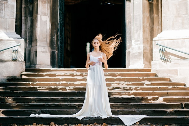 Young romantic elegant girl with flying hair in long white dress posing on the stairs of ancient palace