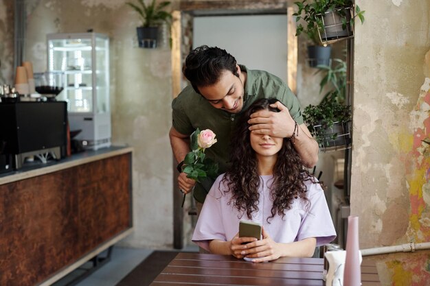 Young romantic man with pink rose covering eyes of his beautiful girlfriend