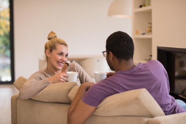 Young romantic multiethnic couple sitting on sofa in front of fireplace at home, looking at each other, talking and drinking coffee