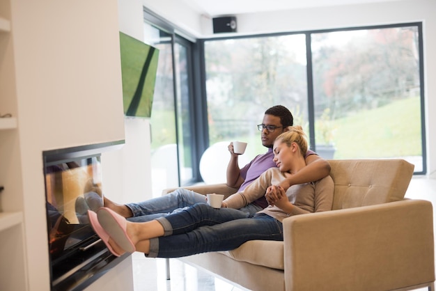 Young romantic multiethnic couple sitting on sofa in front of fireplace at home, looking at each other, talking and drinking coffee