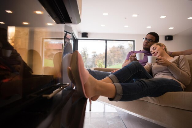 Young romantic multiethnic couple sitting on sofa in front of fireplace at home, looking at each other, talking and drinking coffee