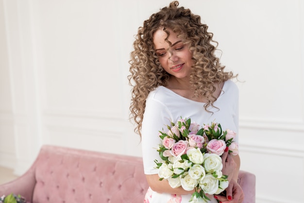 Young romantic woman at home sitting on the pink sofa holding a flowers bouquet and dreaming- Image 