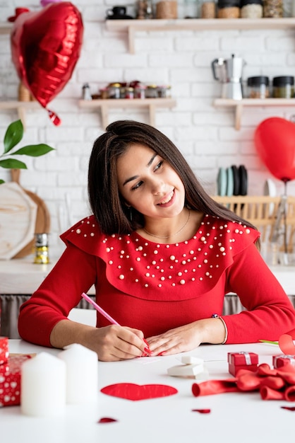 Giovane donna romantica in abito rosso scrivendo lettera d'amore seduto alla cucina decorata