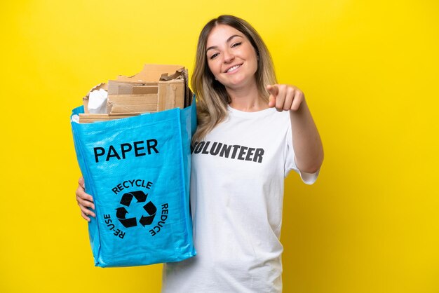 Young Rumanian woman holding a recycling bag full of paper to recycle isolated on yellow background points finger at you with a confident expression