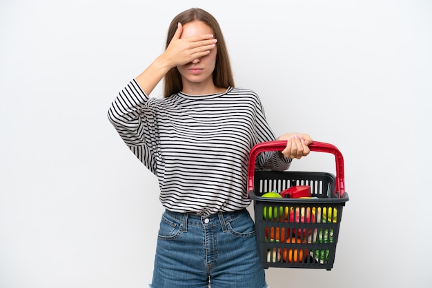 Young Rumanian woman holding a shopping basket full of food isolated on white background covering eyes by hands Do not want to see something