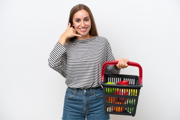 Young Rumanian woman holding a shopping basket full of food isolated on white background making phone gesture Call me back sign