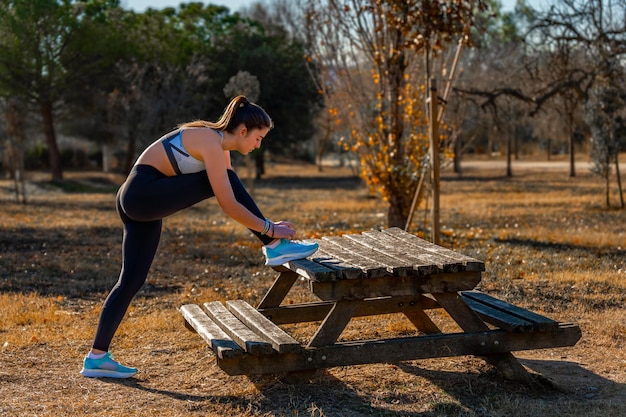 Photo a young runner girl dressed in tight leggings and a top tying the laces of her sneakers resting her foot on a wooden table bench on a sunny morning day in a public mountain park