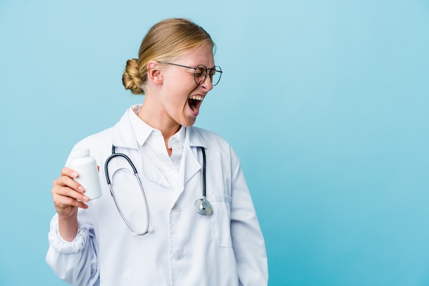 Young russian doctor woman holding pills bottle on blue shouting towards a copy space