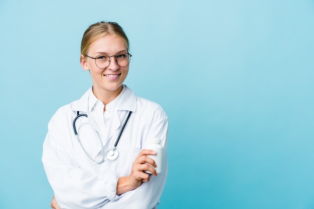 Young russian doctor woman holding pills bottle on blue smiling confident with crossed arms