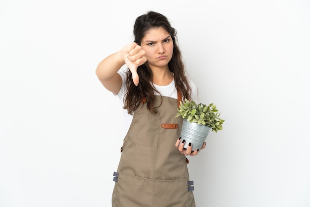 부정적인 표현으로 아래로 엄지를 보여주는 고립 된 식물을 들고 젊은 러시아 정원사 소녀