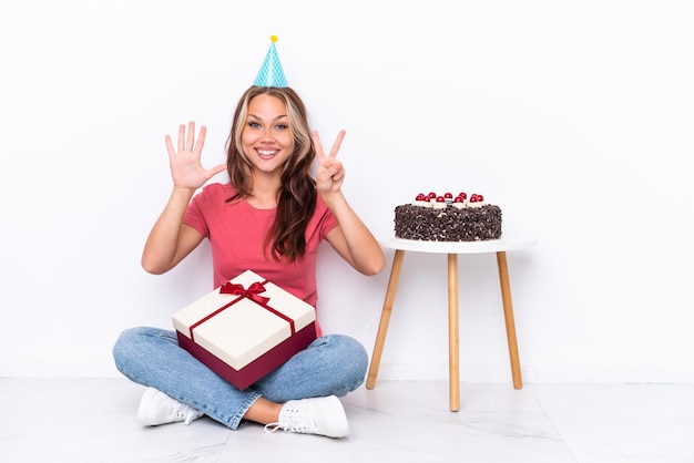 Молодая русская девушка празднует день рождения, сидя на полу, изолированном на белом фоне, считая пальцами семь