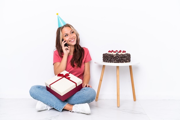 Young Russian girl celebrating a birthday sitting one the floor isolated on white background keeping a conversation with the mobile phone with someone