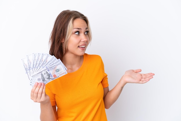 Young Russian girl holding dollars isolated on white background with surprise expression while looking side