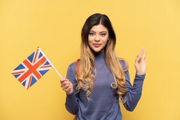 Young Russian girl holding an United Kingdom flag isolated on yellow background showing ok sign with fingers