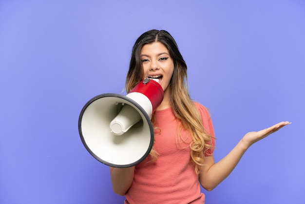 Young Russian girl isolated on blue background shouting through a megaphone and pointing side