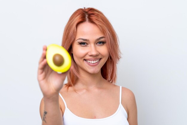 Young Russian girl isolated on white background holding an avocado while smiling Close up portrait