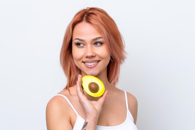 Young Russian girl isolated on white background holding an avocado while smiling Close up portrait