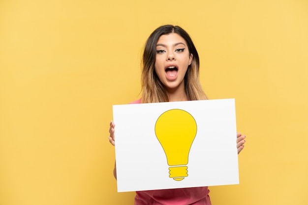 Young Russian girl isolated on yellow background holding a placard with bulb icon with happy expression