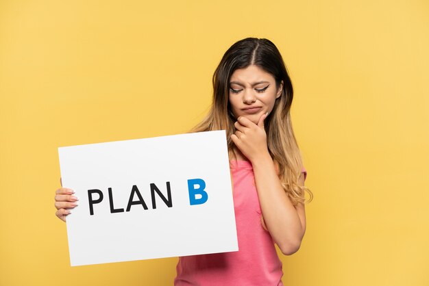 Young Russian girl isolated on yellow background holding a placard with the message PLAN B and thinking