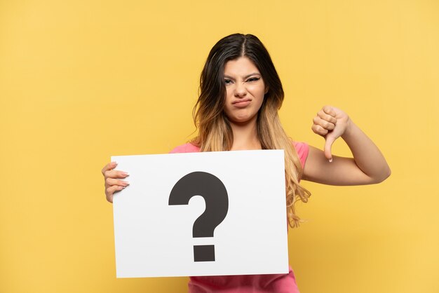 Young Russian girl isolated on yellow background holding a placard with question mark symbol and doing bad signal