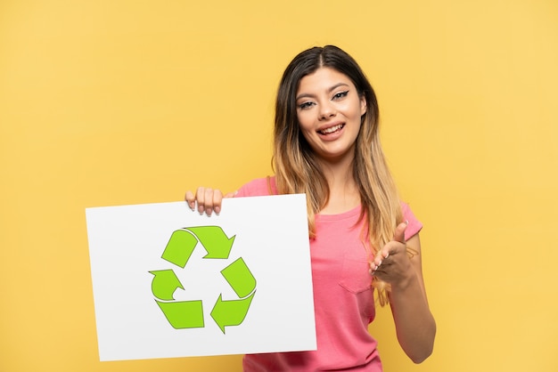 Young Russian girl isolated on yellow background holding a placard with recycle icon making a deal