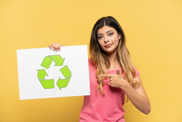 Young Russian girl isolated on yellow background holding a placard with recycle icon and  pointing it