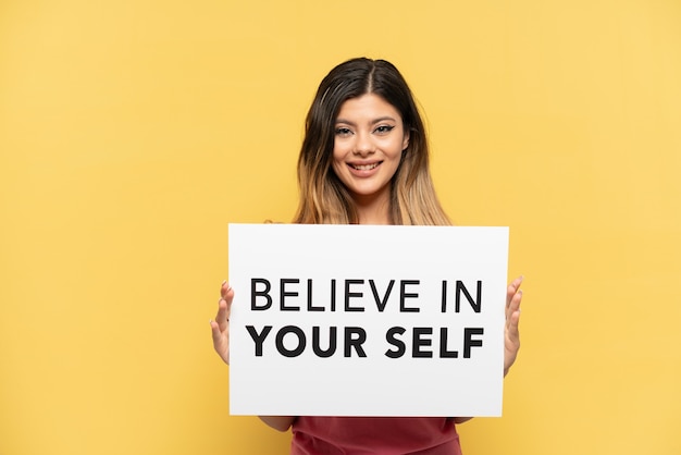Young Russian girl isolated on yellow background holding a placard with text Believe In Your Self with happy expression