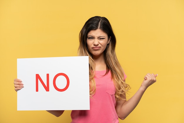 Young Russian girl isolated on yellow background holding a placard with text NO and angry