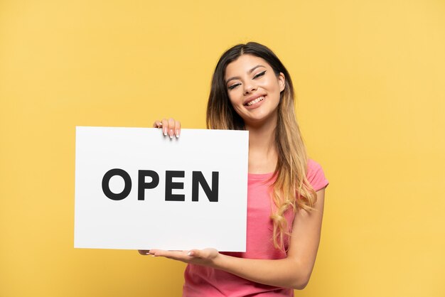 Young Russian girl isolated on yellow background holding a placard with text OPEN with happy expression