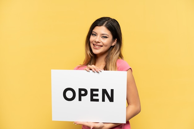 Young Russian girl isolated on yellow background holding a placard with text OPEN with happy expression