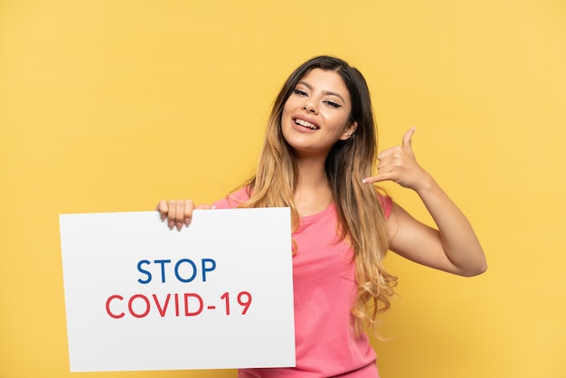 黄色の背景に孤立した若いロシアの女の子は、テキストStop Covid 19のプラカードを保持し、電話ジェスチャーをしています