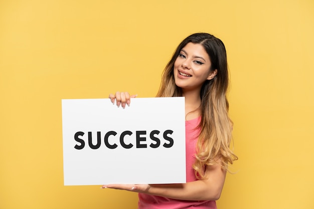 Young Russian girl isolated on yellow background holding a placard with text SUCCESS with happy expression