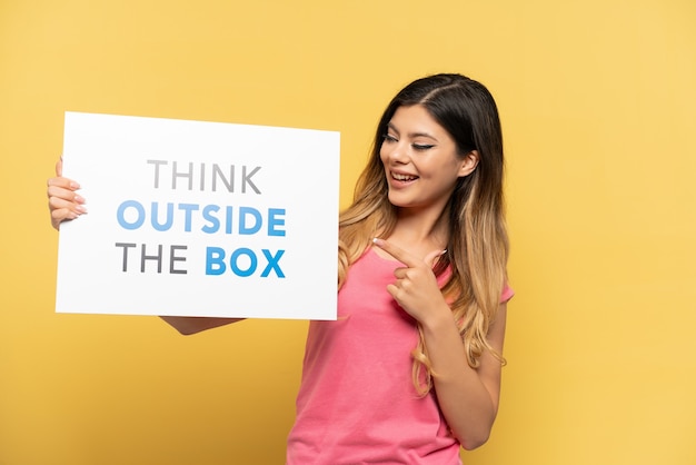 노란색 배경에 격리된 러시아 소녀는 Think Outside the Box라는 문구가 적힌 현수막을 들고 그것을 가리키고 있습니다.