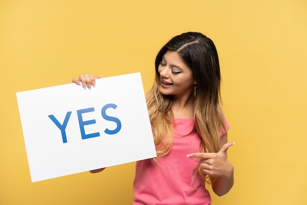 Young Russian girl isolated on yellow background holding a placard with text YES and pointing it