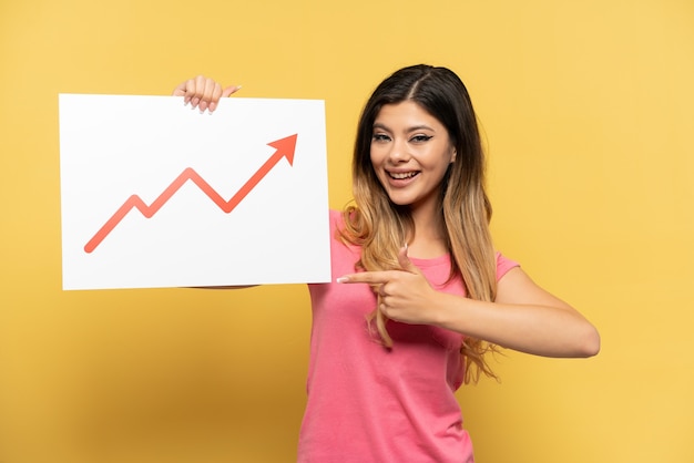 Young Russian girl isolated on yellow background holding a sign with a growing statistics arrow symbol and  pointing it