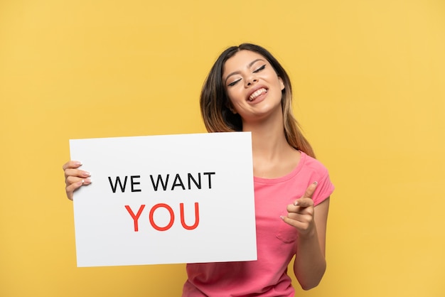 Young Russian girl isolated on yellow background holding We Want You board and pointing to the front