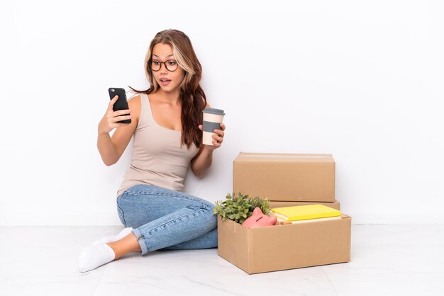 Young Russian girl sitting on the floor isolated on white background holding coffee to take away and a mobile