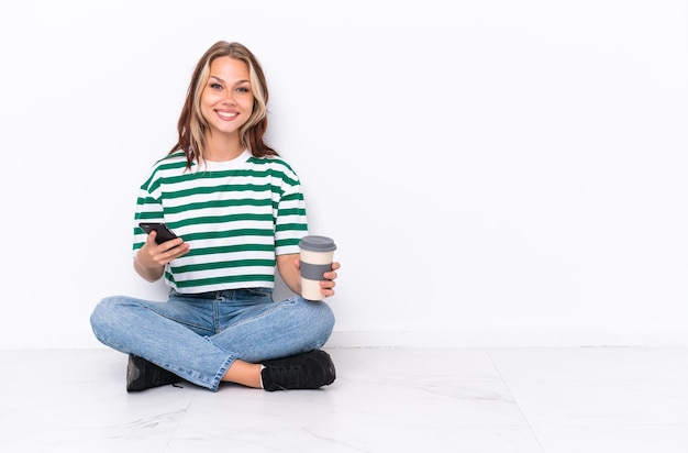 持ち帰り用のコーヒーと携帯電話を保持している白い背景で隔離の床に座っている若いロシアの女の子