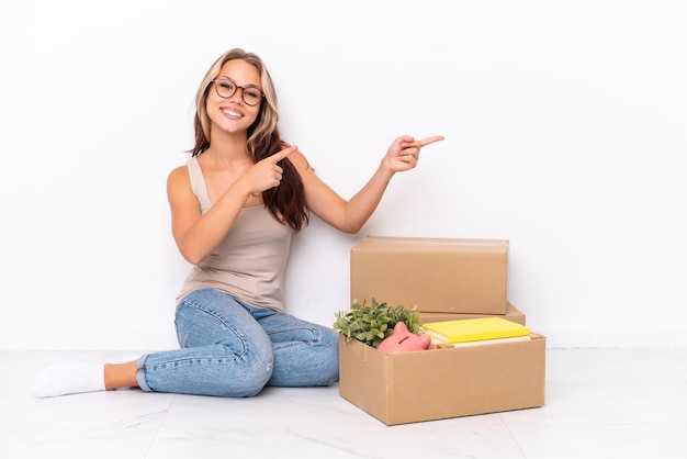 Young Russian girl sitting on the floor isolated on white background pointing finger to the side and presenting a product