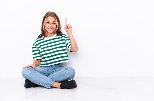 Giovane ragazza russa seduta sul pavimento isolata su sfondo bianco che punta con il dito indice una grande idea