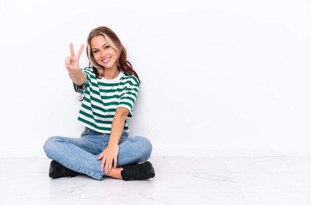 Young Russian girl sitting on the floor isolated on white background smiling and showing victory sign