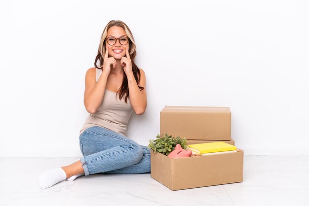 Young Russian girl sitting on the floor isolated on white background smiling with a happy and pleasant expression