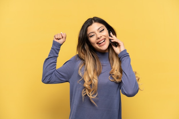 Young Russian girl using mobile phone isolated on yellow background celebrating a victory