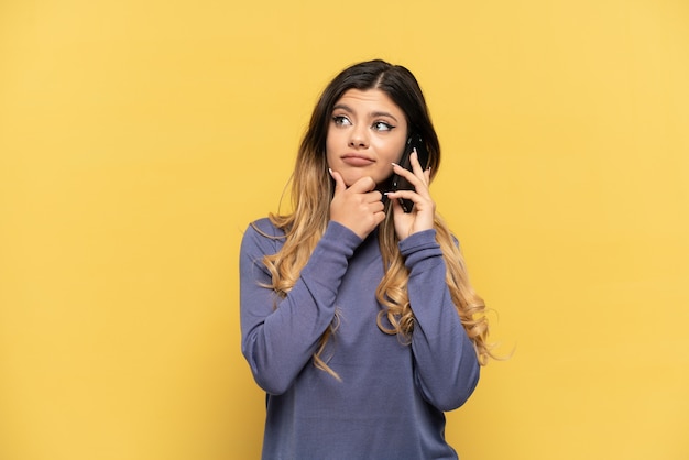 Young Russian girl using mobile phone isolated on yellow background looking up while smiling